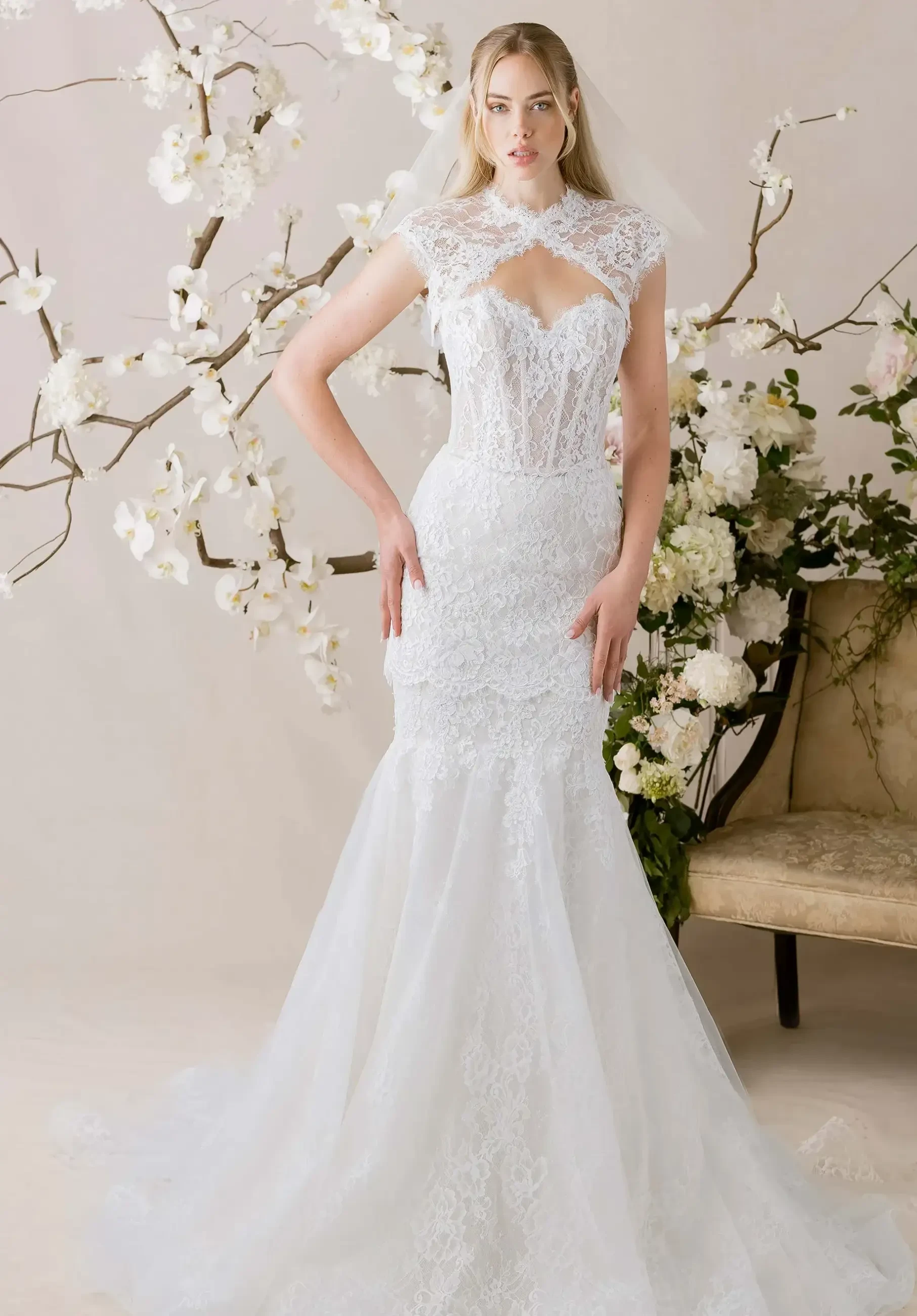 A model wearing an elegant white wedding dress with lace detailing and a fitted silhouette, standing in front of floral decor.