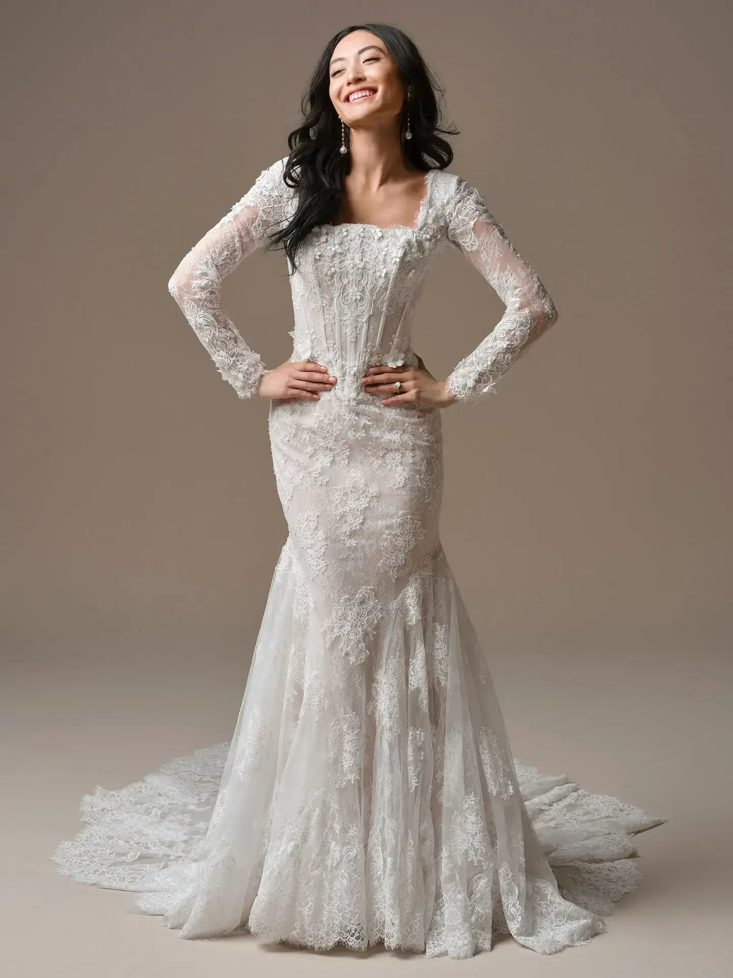 A woman wearing a long-sleeve, lace wedding dress, smiling and posing confidently.
