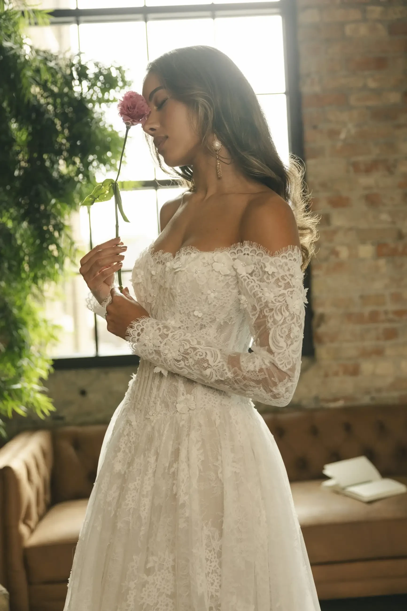 A woman in a white lace wedding dress holds a pink flower near her face while standing indoors next to a large window.