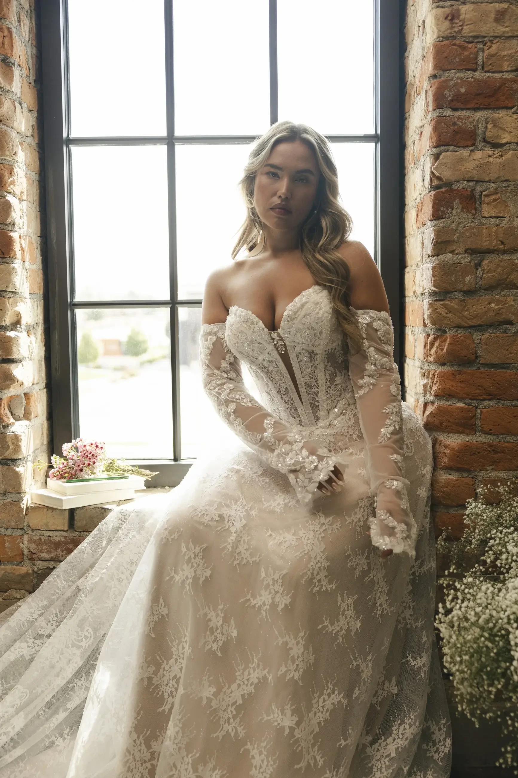 A woman in a bridal gown sitting by a large window with exposed brick walls. The dress has intricate lace details and long sleeves, and she looks thoughtfully at the camera.