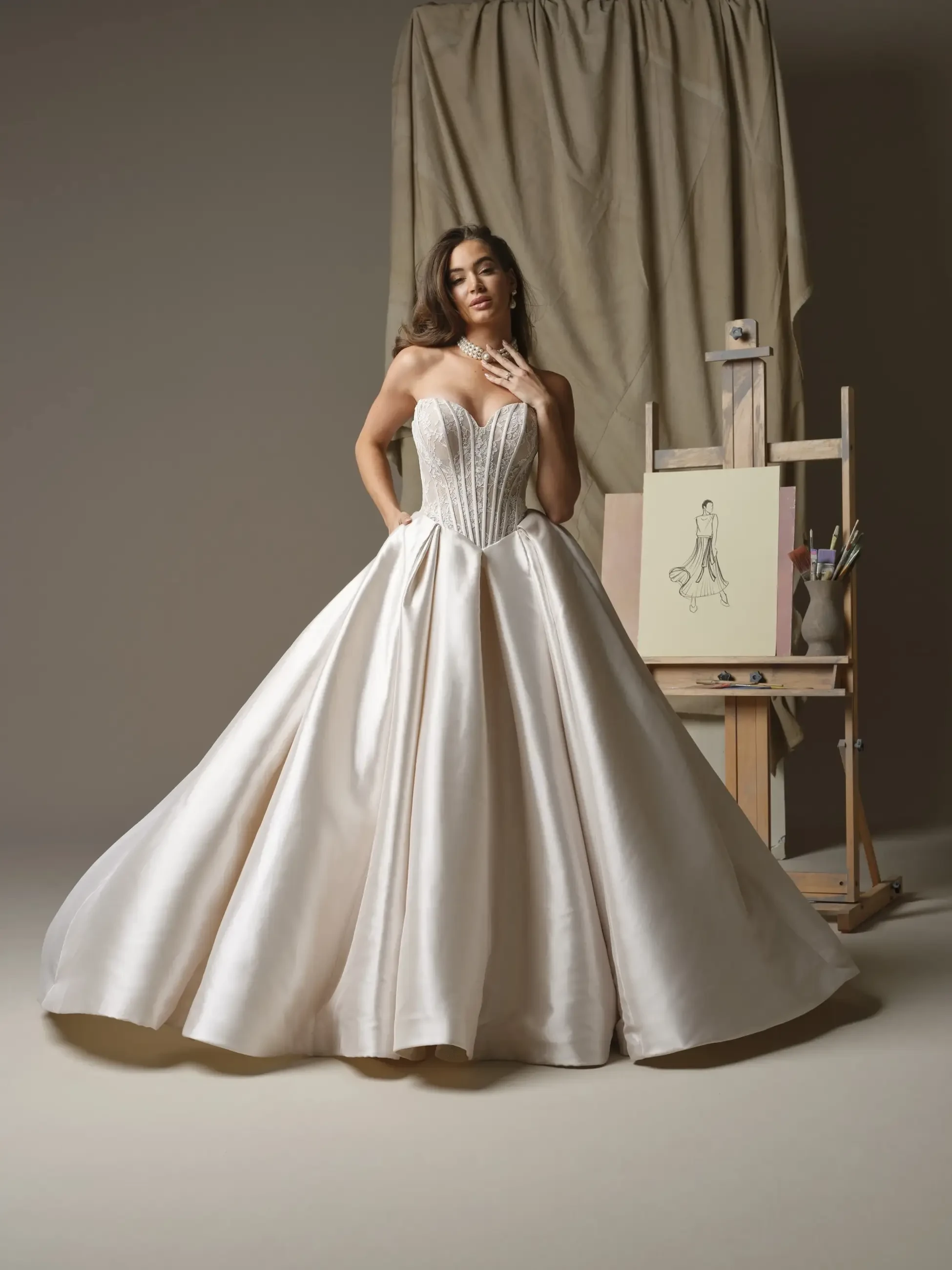 A woman in a strapless, elegant white gown stands gracefully, posing in front of an easel with a sketch of a dress. The background features draped fabric, enhancing the artistic ambiance.