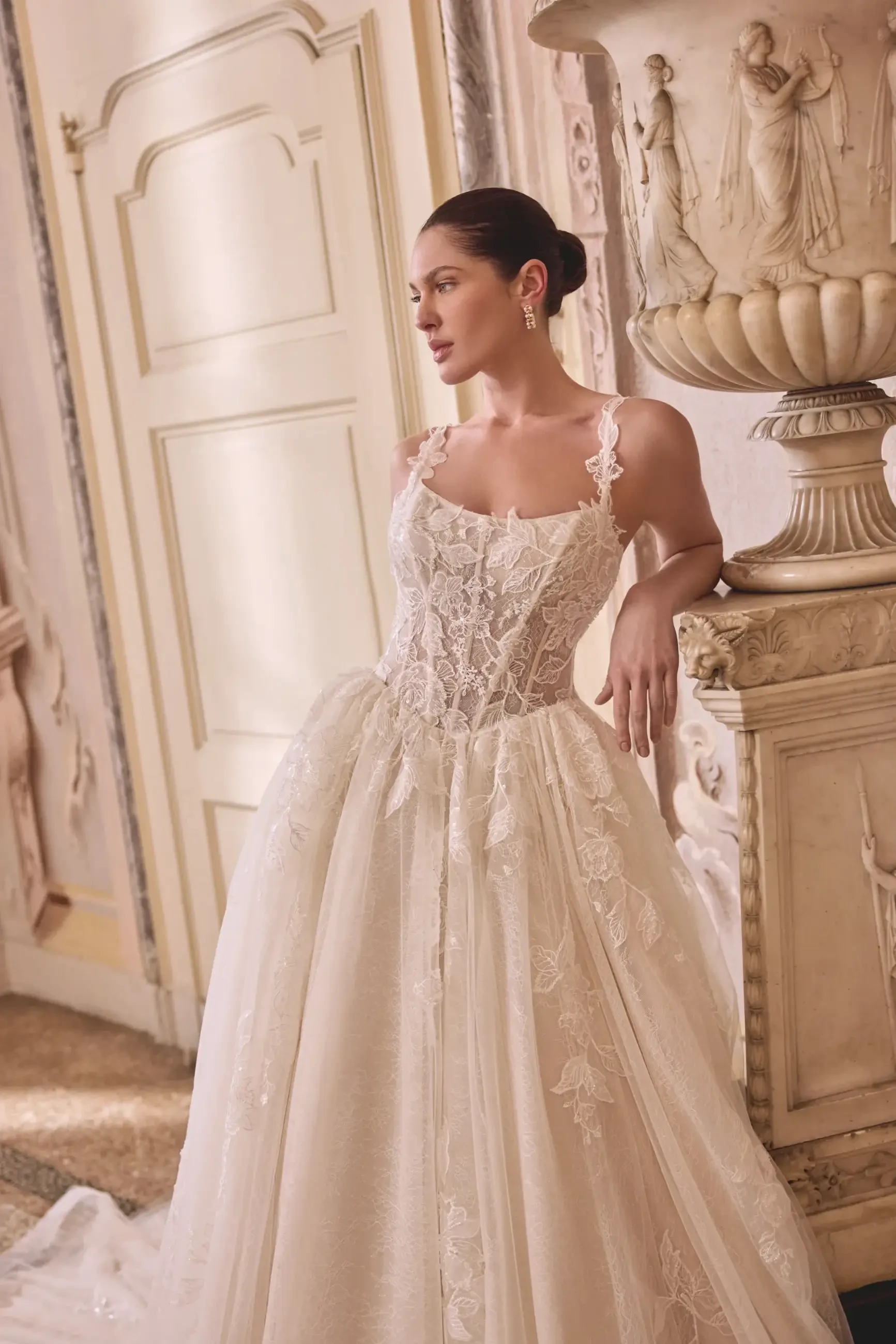 A model wearing an elegant wedding dress with lace detailing, posing in a luxurious indoor setting.