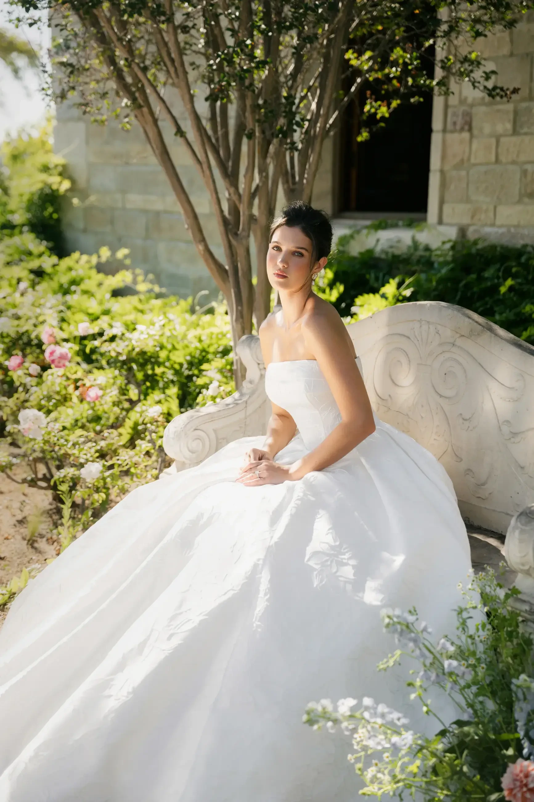 A young woman in a strapless white wedding gown sits on a decorative bench surrounded by greenery and flowers.