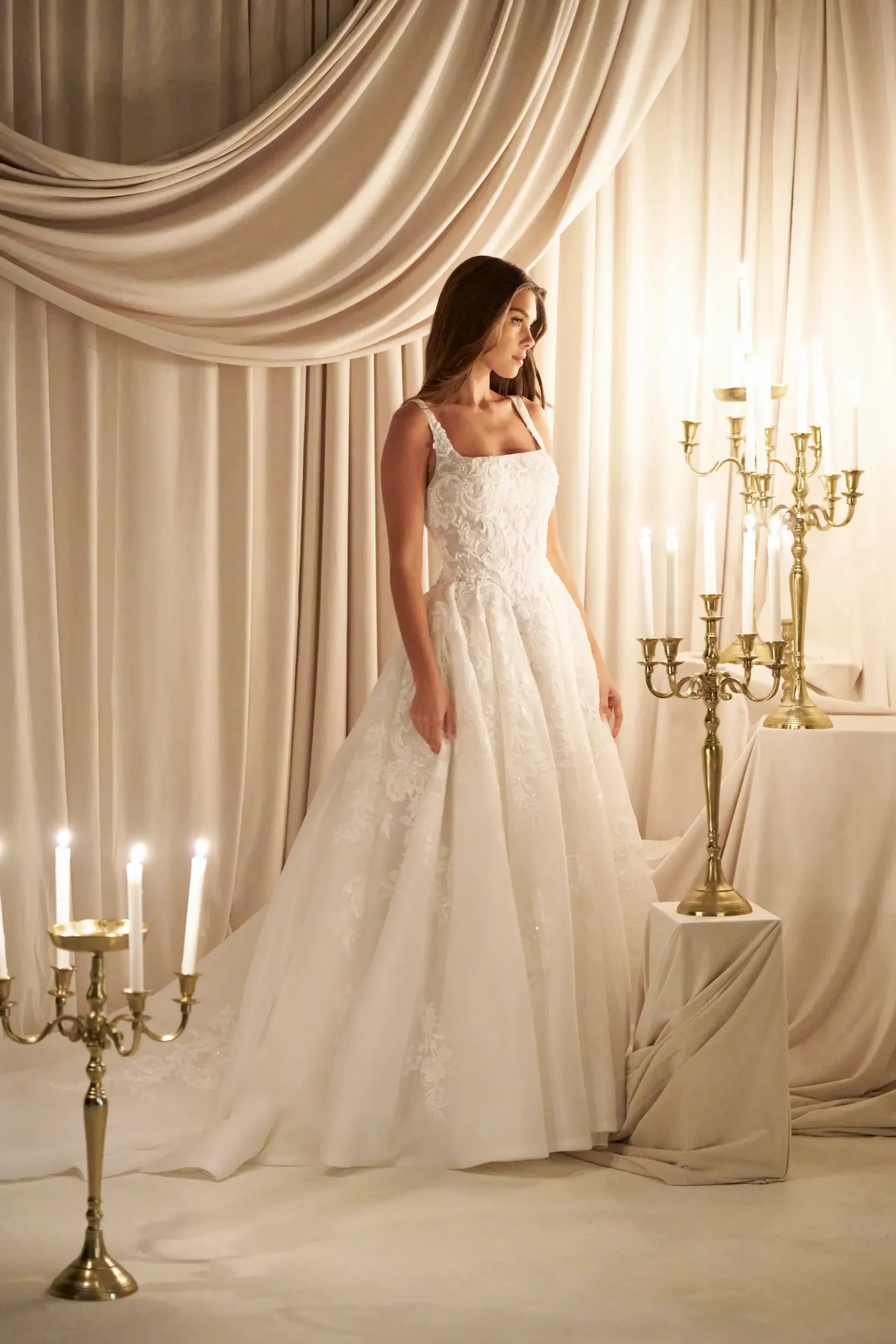 A woman in a white wedding dress standing near lit candelabras, with draped fabric in the background.