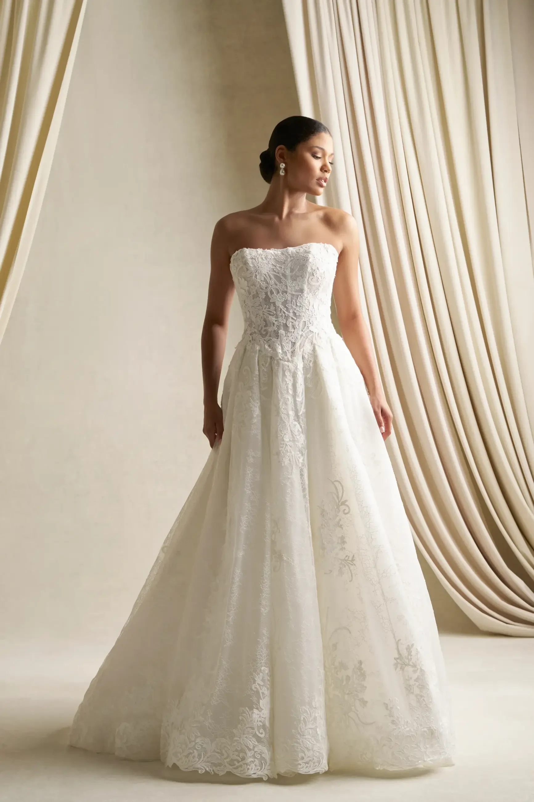 A woman wearing a strapless white wedding dress stands gracefully against a backdrop of soft, draped fabric.